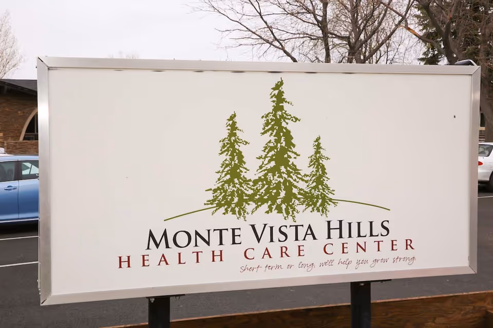 Outdoor sign for Monte Vista Hills Health Care Center featuring three green pine trees and the tagline 'Short term or long, we'll help you grow strong' with a parking lot and trees in the background.