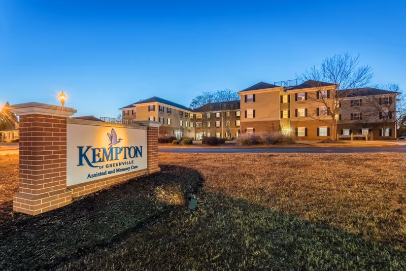 Exterior view of a multi-story assisted living facility building during dusk with lights on inside. In the foreground, there is a brick sign that reads 'KEMPTON of GREENVILLE Assisted and Memory Care'. The sky is clear and blue, and the surrounding area has grass and some trees without leaves.