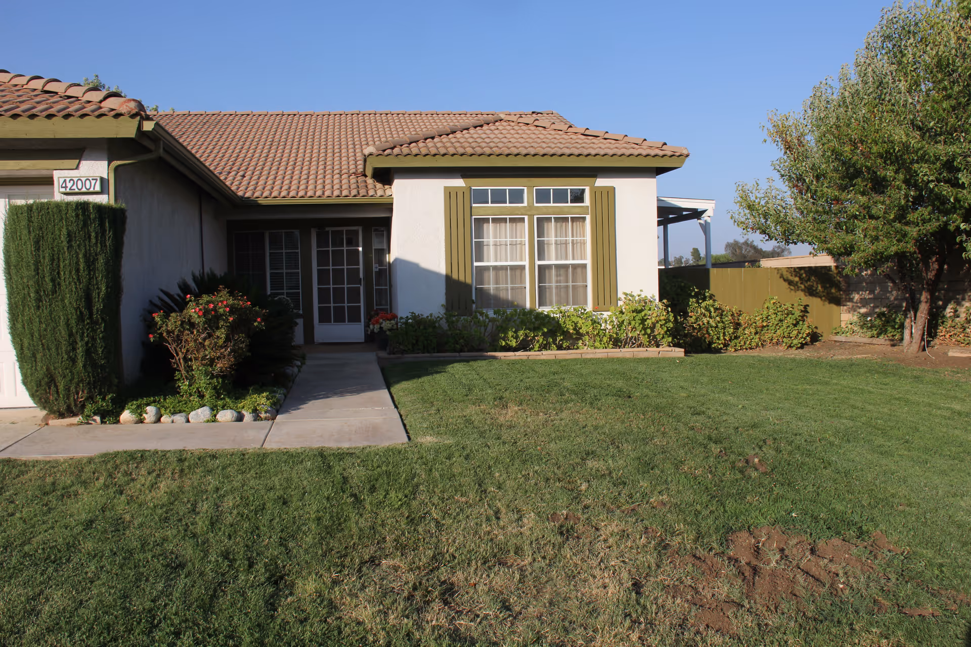 Front exterior of a single-story house with a tiled roof, porch entry, shrubs and a green lawn.