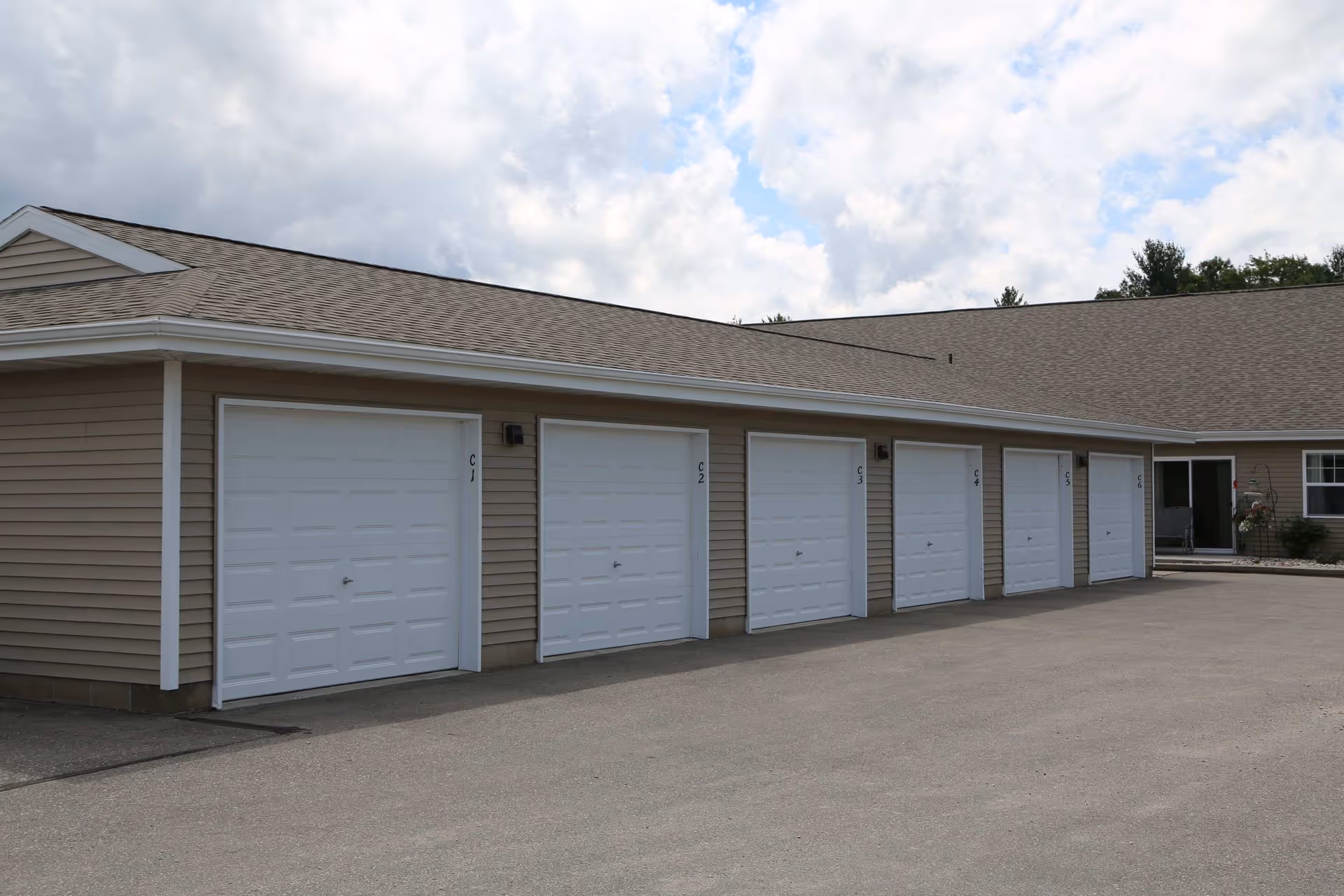 A row of six closed white garage doors on a beige building with a sloped roof under a partly cloudy sky. The garages are numbered C1 to C6. There is a paved driveway in front of the garages and a small entrance area with a door and window at the far right.