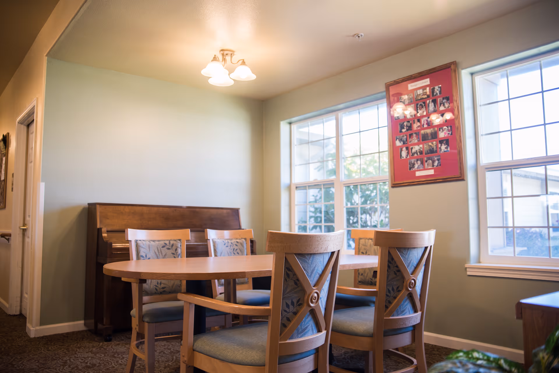 A cozy dining area with a wooden oval table surrounded by six chairs with patterned cushions. Behind the table is an upright piano against a light green wall. Two large windows let in natural light, and a framed collage of photos hangs on the wall between the windows. A ceiling light fixture with three bulbs illuminates the room.