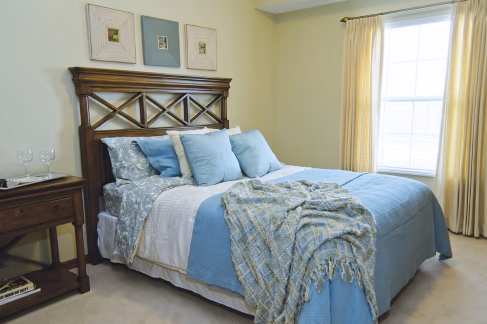 A neatly made bed with a wooden headboard, adorned with blue and white bedding and multiple pillows. A blue and beige patterned throw blanket is draped over the foot of the bed. To the left of the bed is a wooden nightstand with two empty wine glasses and a book on the lower shelf. The room has light-colored walls and a large window with beige curtains allowing natural light to enter.