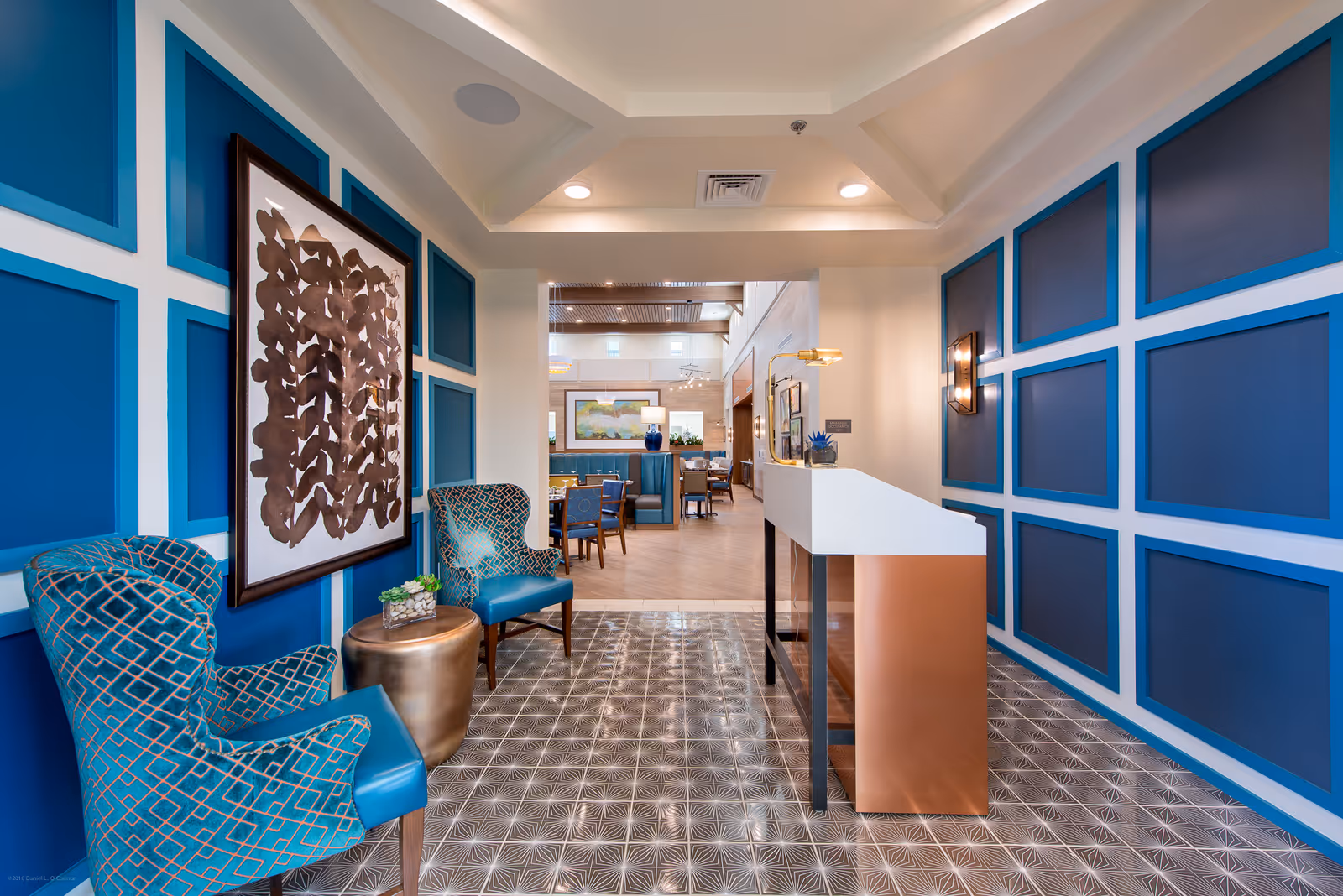 A stylish interior hallway with blue paneled walls and patterned floor tiles. Two blue upholstered chairs with geometric patterns flank a small round bronze table with a plant on it. A large abstract artwork hangs on the left wall. The hallway leads into a dining area with tables and chairs, visible in the background.