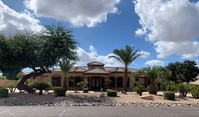 Single-story stucco building with a tiled roof, arched entrance, and desert landscaping with palm trees under a partly cloudy sky.