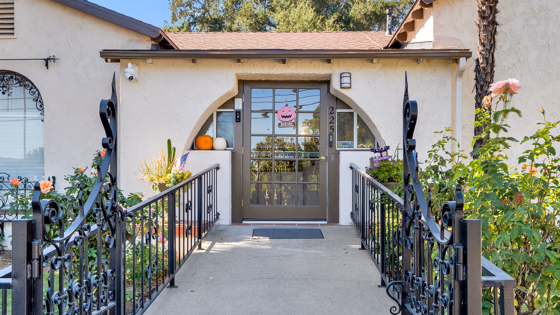Front entrance of a building with a brown door featuring glass panes, flanked by two small white walls with pumpkins and plants on top. There is a black wrought iron gate and railing leading up to the door, with rose bushes and other greenery on either side. A pink Halloween decoration with a jack-o'-lantern face hangs on the door, and the building has a light beige stucco exterior with a brown shingled roof.