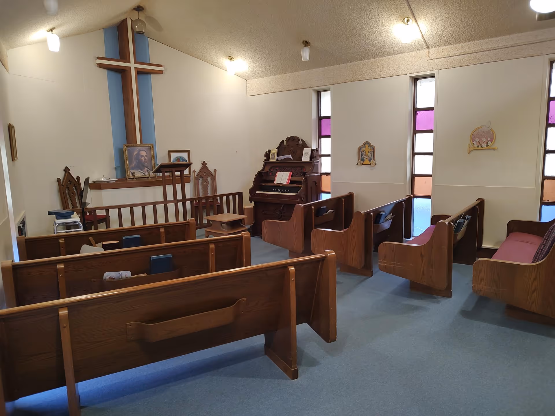 Interior of a small chapel with wooden pews, a large wooden cross on the wall, a framed picture of Jesus, an organ, and stained glass windows with purple and orange panels.
