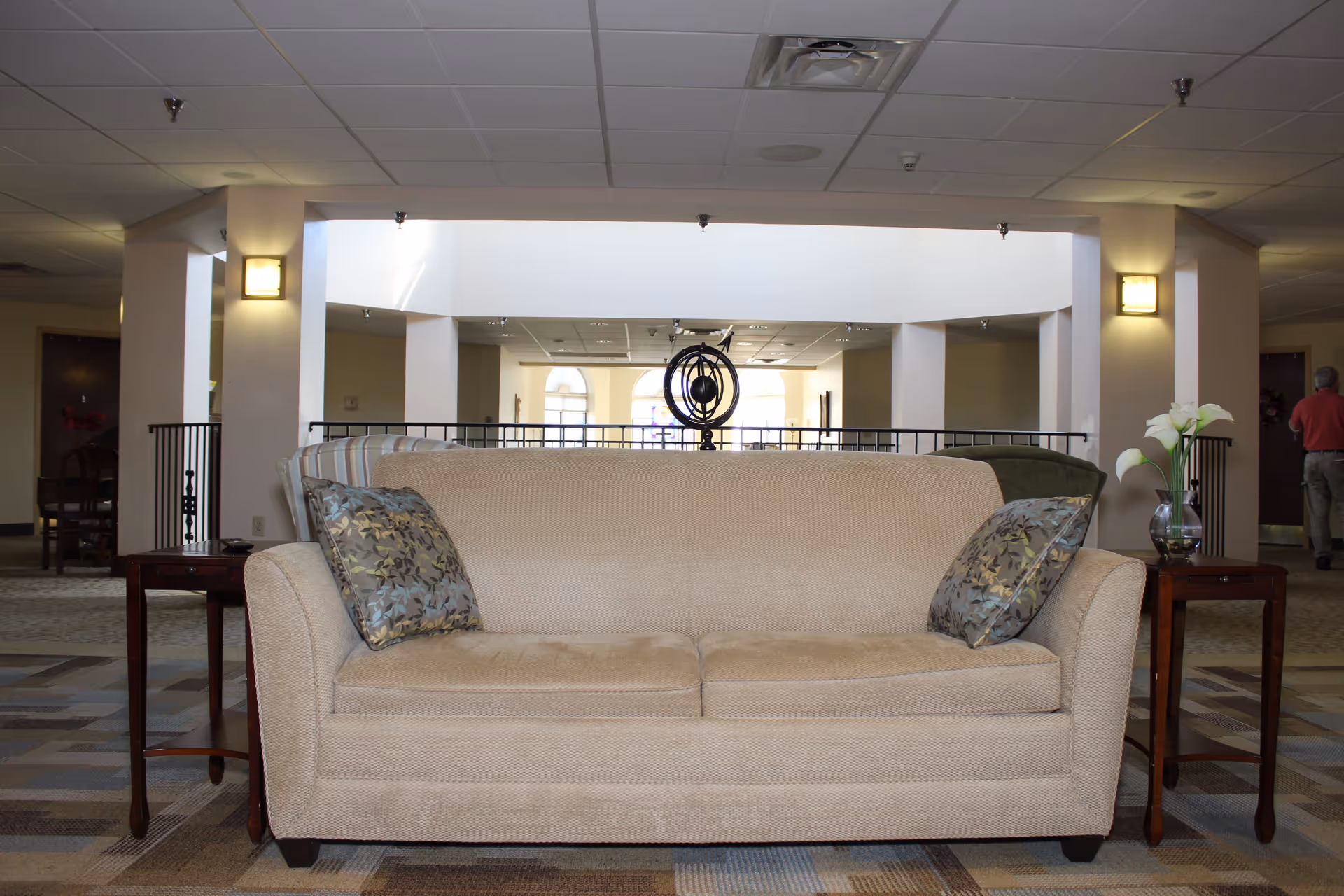 A beige sofa with two patterned pillows is centered in a spacious, well-lit common area. On each side of the sofa is a wooden side table, one with a glass vase holding white flowers. The background shows a railing, columns, and a ceiling with recessed lighting. A person is walking away in the distance on the right side.