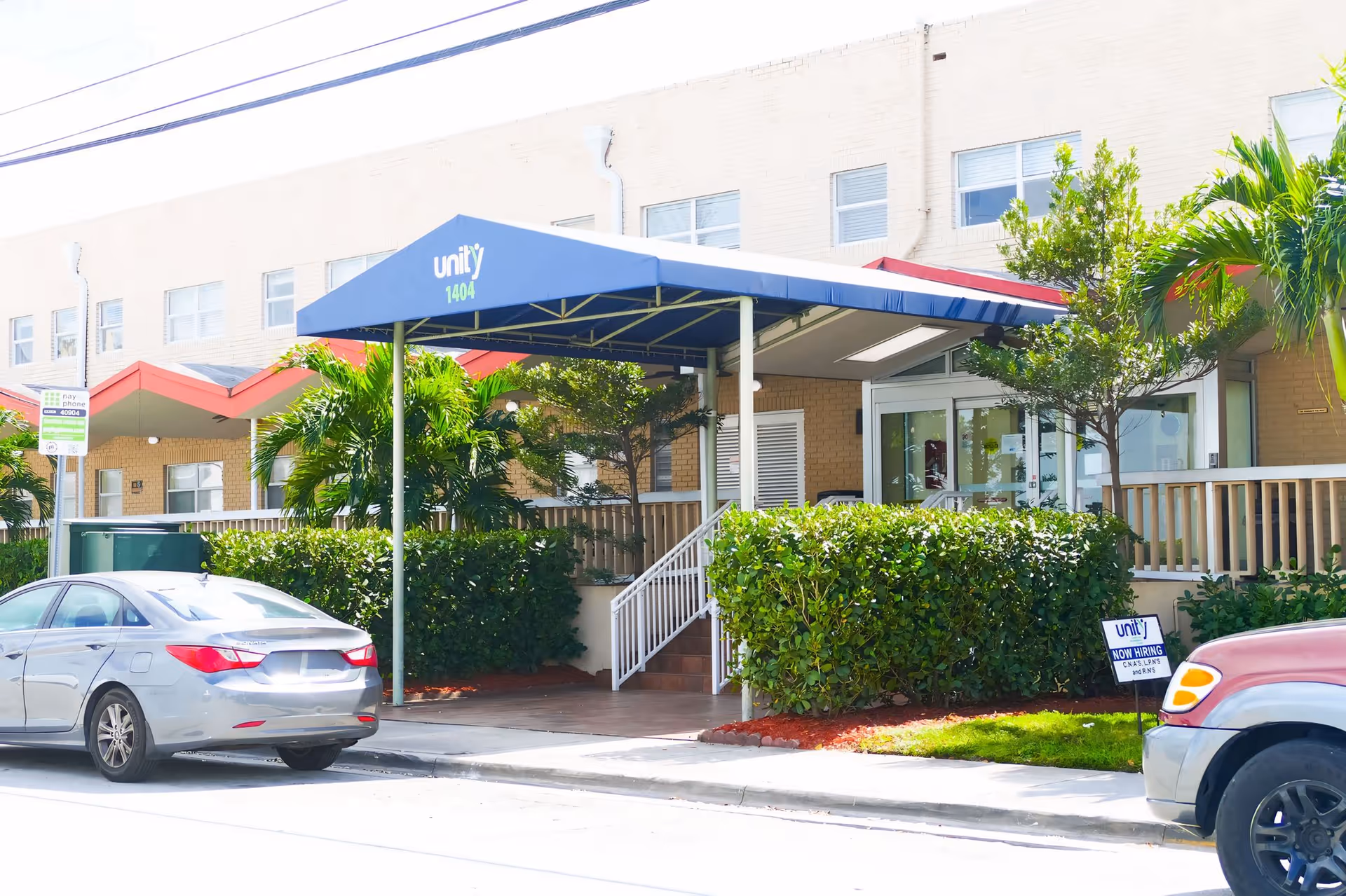 Front entrance of Unity Health and Rehabilitation Center showing a blue awning reading 'unity 1404', a ramp and steps, hedges, and parked cars.
