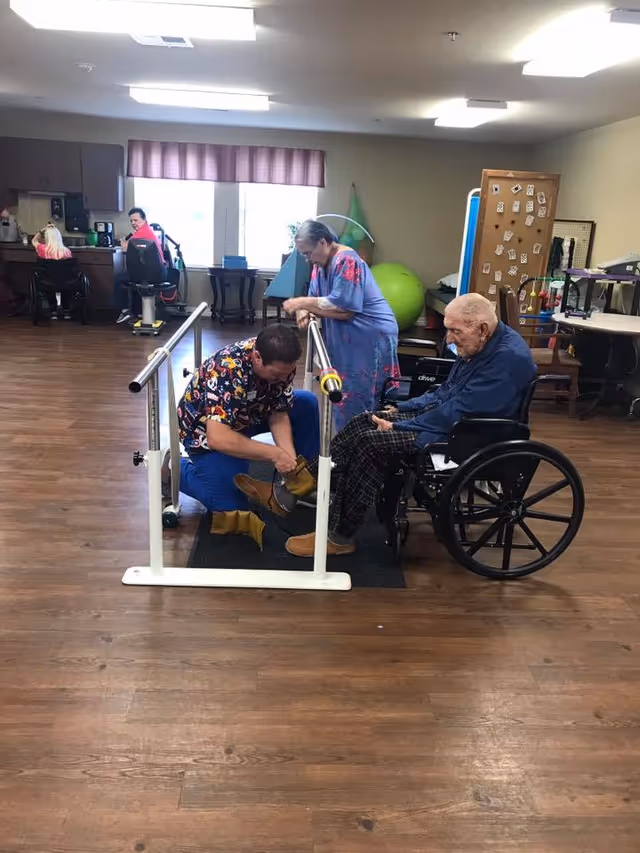 Staff and residents in a senior living activity/rehab room, with a caregiver fitting boots on a seated man in a wheelchair at parallel bars.