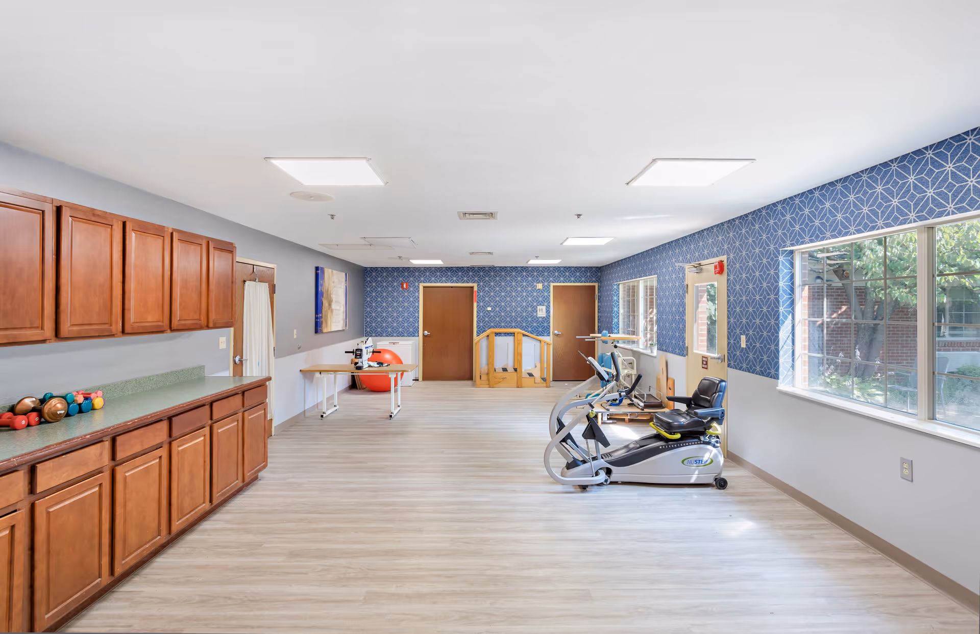 Bright rehabilitation/activity room with wooden cabinets on the left, exercise equipment and therapy balls on the right, and large windows letting in natural light.