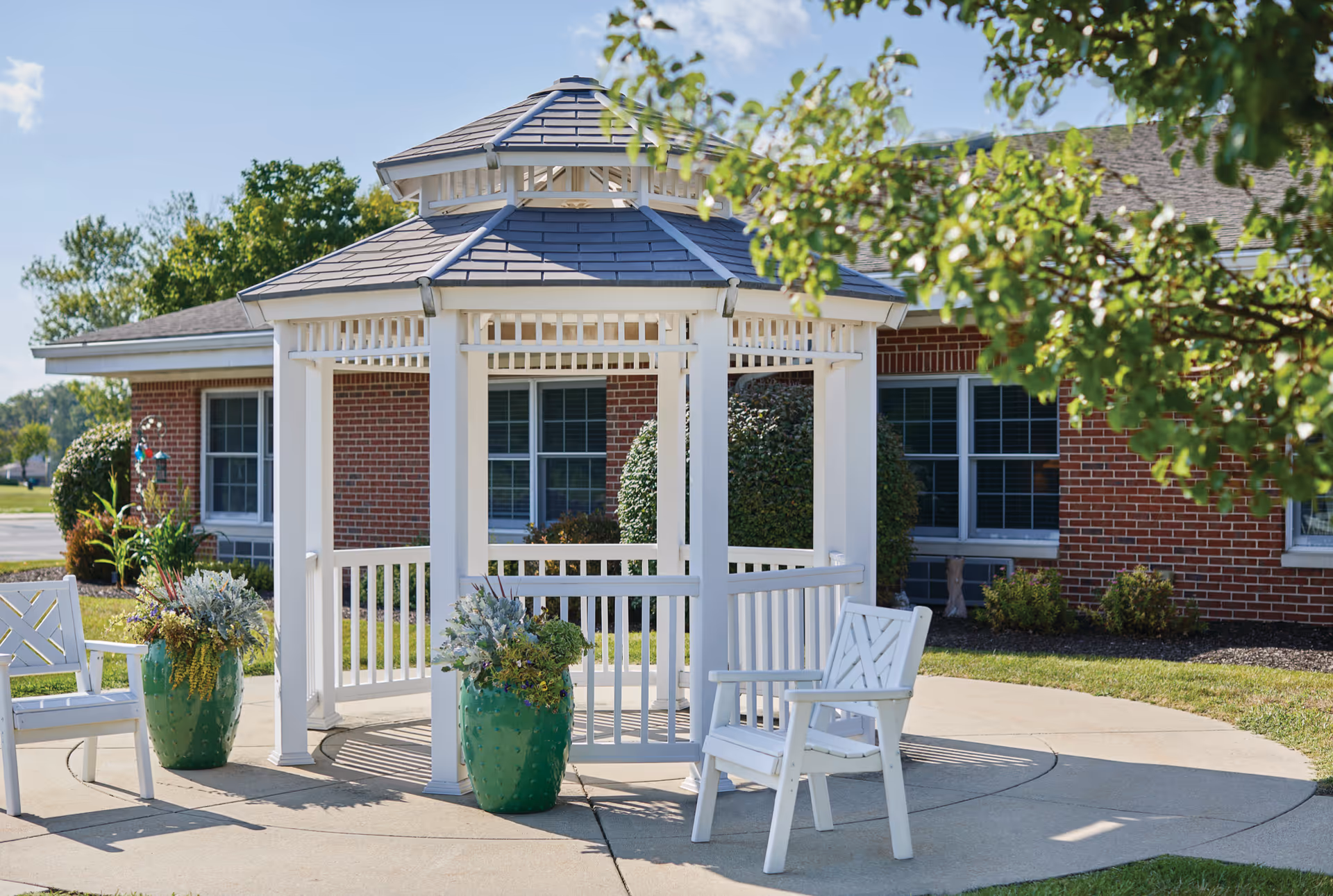 White wooden gazebo with a gray roof situated on a concrete patio in front of a red brick building. Two white wooden chairs and two large green planters with plants are placed around the gazebo. Green trees and shrubs surround the area under a clear blue sky.