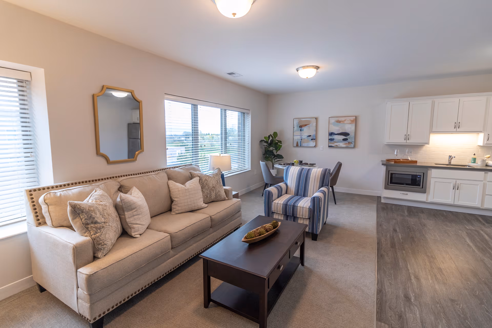 A bright and cozy living room area with a beige sofa adorned with multiple pillows, a striped armchair, and a dark wooden coffee table with decorative items. The room features large windows with blinds, a wall mirror, two abstract paintings, a small dining table with chairs, and a kitchen area with white cabinets, a microwave, and a sink.