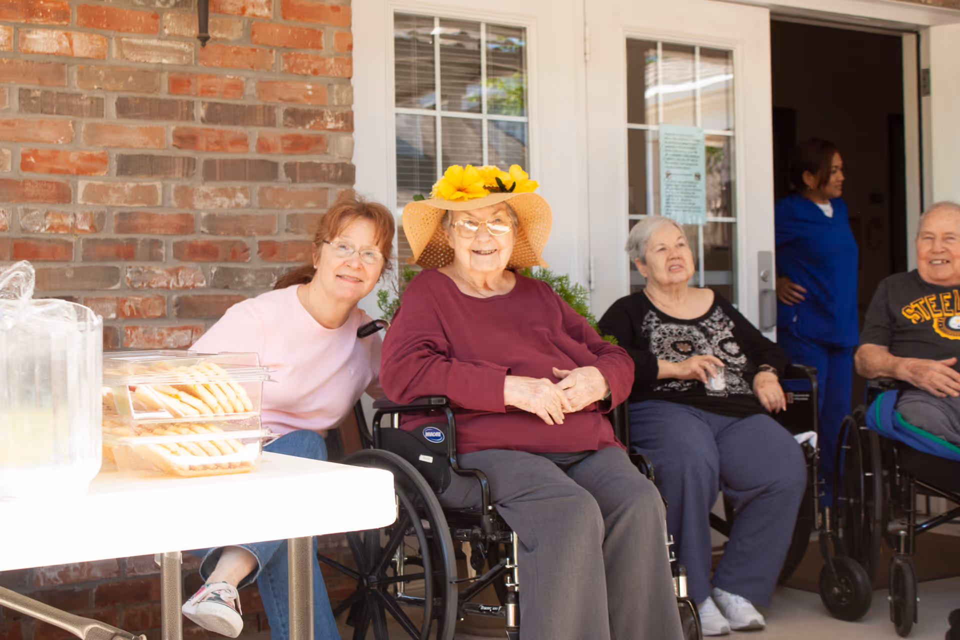 A group of elderly people and a caregiver sitting outside a building. One elderly woman in a wheelchair is wearing a large straw hat with yellow flowers and a maroon sweater. Another elderly woman and a man in wheelchairs are sitting nearby, along with a caregiver in blue scrubs standing in the background. A woman in a pink shirt is sitting next to the elderly woman with the hat. A table with a pitcher of water and a container of crackers is in the foreground.