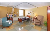 A well-lit room with a hospital-style bed in the center, a walker positioned next to the bed, a blue armchair on the left, a beige recliner on the right, a wooden cabinet, and a green laundry basket. The walls are painted light yellow, and there are two windows letting in natural light.