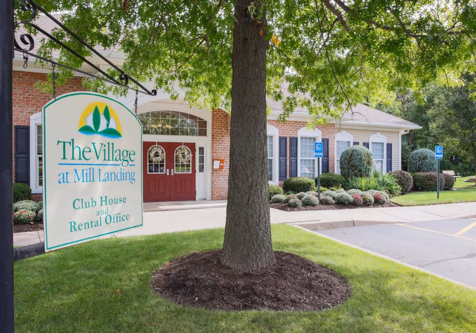 Exterior view of The Village at Mill Landing clubhouse and rental office building with a large tree in the foreground and a sign displaying the facility name. The building has red double doors, brick and siding walls, and landscaped bushes. There are two handicap parking signs visible near the parking lot.