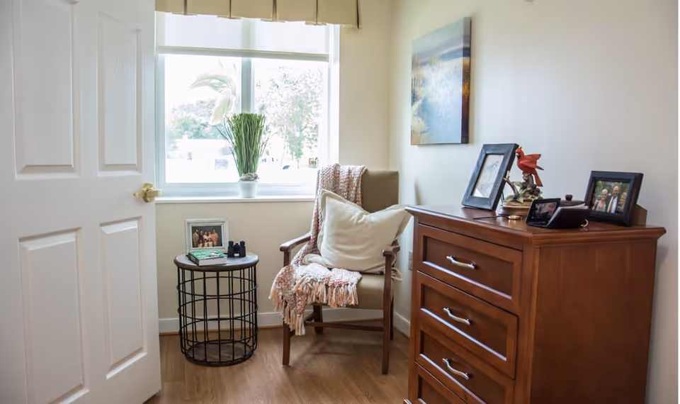 A cozy corner of a room featuring a wooden armchair with a white pillow and a pink throw blanket. Next to the chair is a small round black wireframe side table with a framed photo, a book, and binoculars on it. A window with a potted plant on the sill lets in natural light. On the right side, there is a wooden dresser with three drawers, decorated with framed photos, a red bird figurine, and a small lamp. A landscape painting hangs on the wall above the dresser.