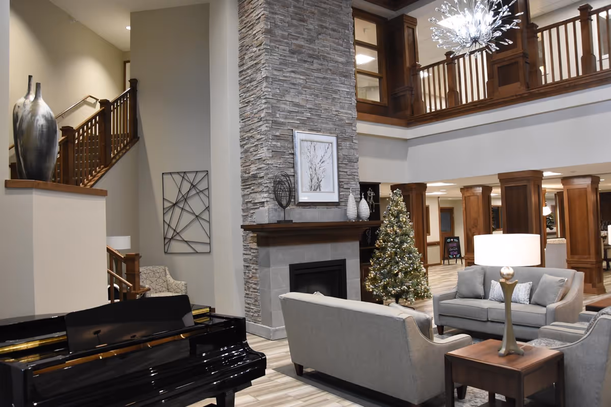A spacious and elegant living room area in a senior living facility featuring a black grand piano, a stone fireplace with decorative items on the mantel, a decorated Christmas tree, and comfortable gray sofas arranged around a wooden side table with a lamp. The room has wooden railings on the upper level and a modern chandelier hanging from the ceiling.