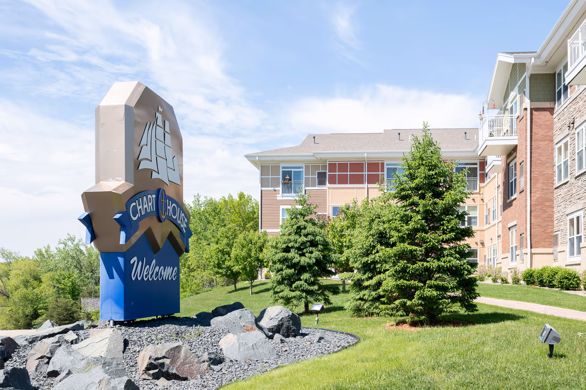 Outdoor view of a senior living facility with a large sign that reads 'Chart House Welcome' surrounded by rocks and greenery. The building is visible in the background with multiple windows and balconies, and there are several trees and a well-maintained lawn in the foreground under a partly cloudy sky.