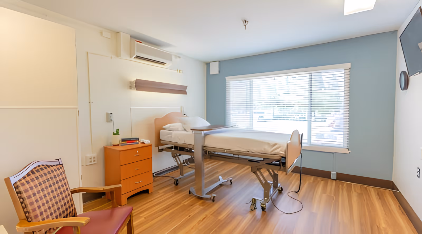 A bright nursing facility room with a hospital bed, a wooden bedside table with books and a plant, a wooden chair with a patterned cushion, a large window with blinds, and a wall-mounted TV.