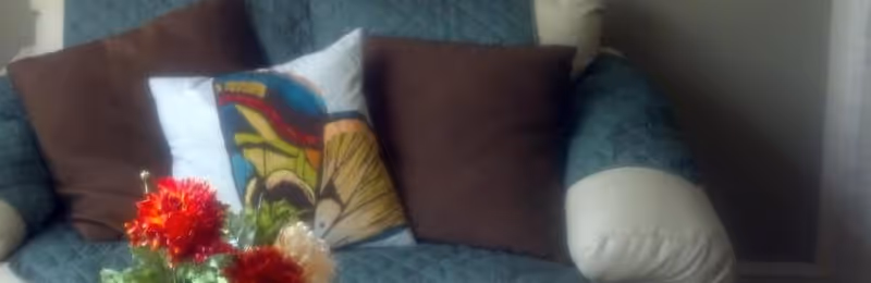 Close-up view of a cushioned armchair with a quilted teal cover, adorned with two brown pillows and one decorative pillow featuring a colorful abstract design. In front of the chair, there is a bouquet of red and white flowers.