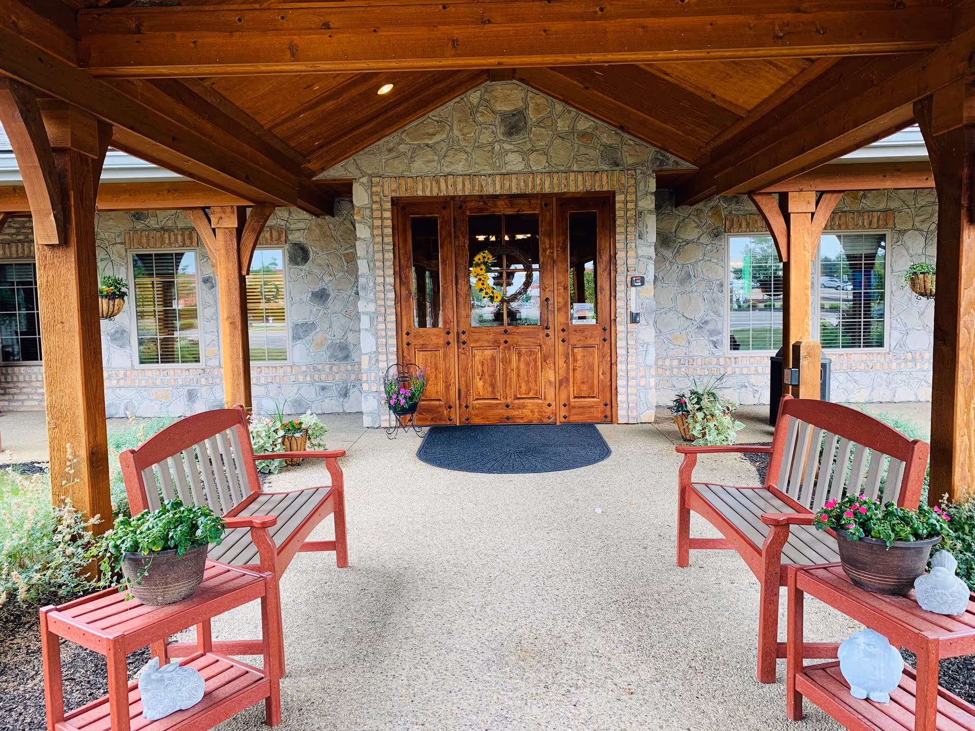 Covered entrance area of The Inn at Winchester Trail featuring a wooden ceiling with beams, stone walls, and a double wooden door decorated with a sunflower wreath. Two red benches with potted plants on small tables are placed on either side of the entrance.