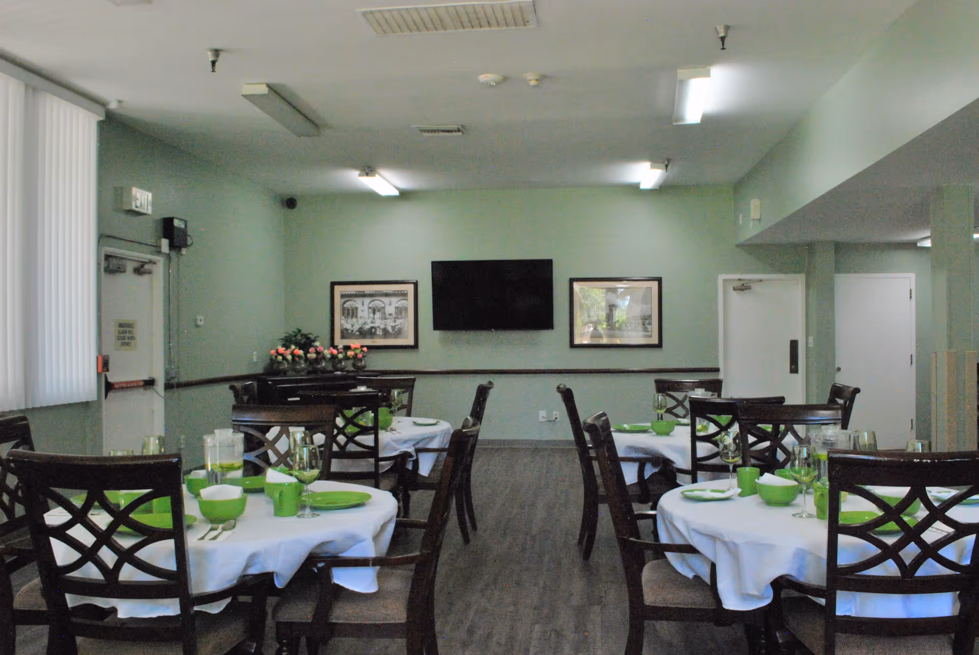 Dining room with round tables covered in white tablecloths, set with green plates, bowls, cups, and glassware. Dark wooden chairs surround the tables. The room has light green walls, a wall-mounted flat screen TV, two framed pictures, and a piano with a flower arrangement on top. There are exit doors and fluorescent ceiling lights.