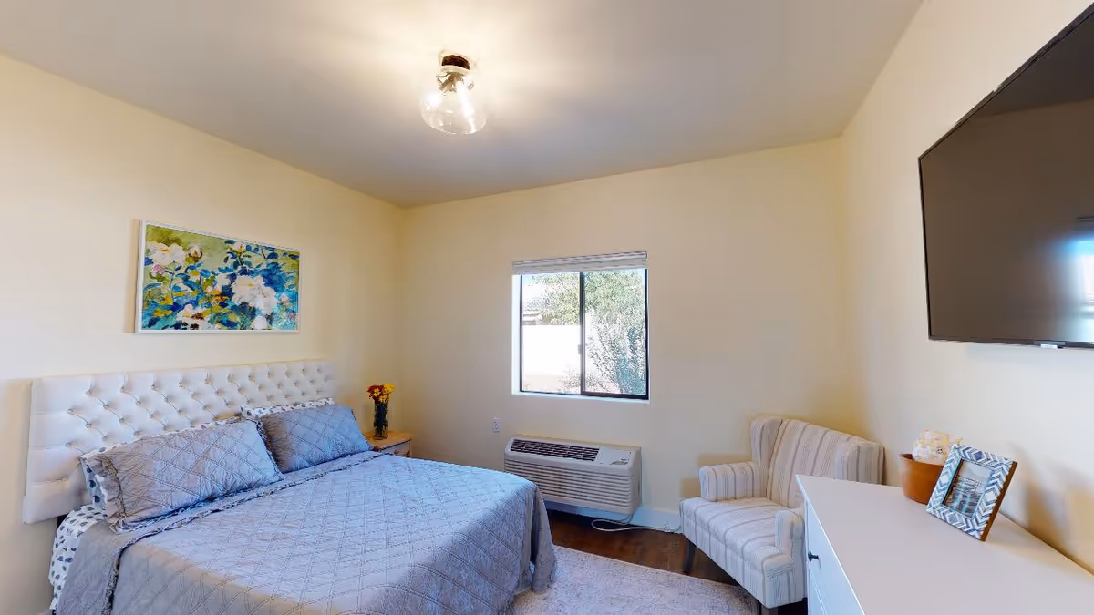 A tidy bedroom with a made bed and tufted headboard, armchair, wall-mounted TV, window, and dresser with decor.