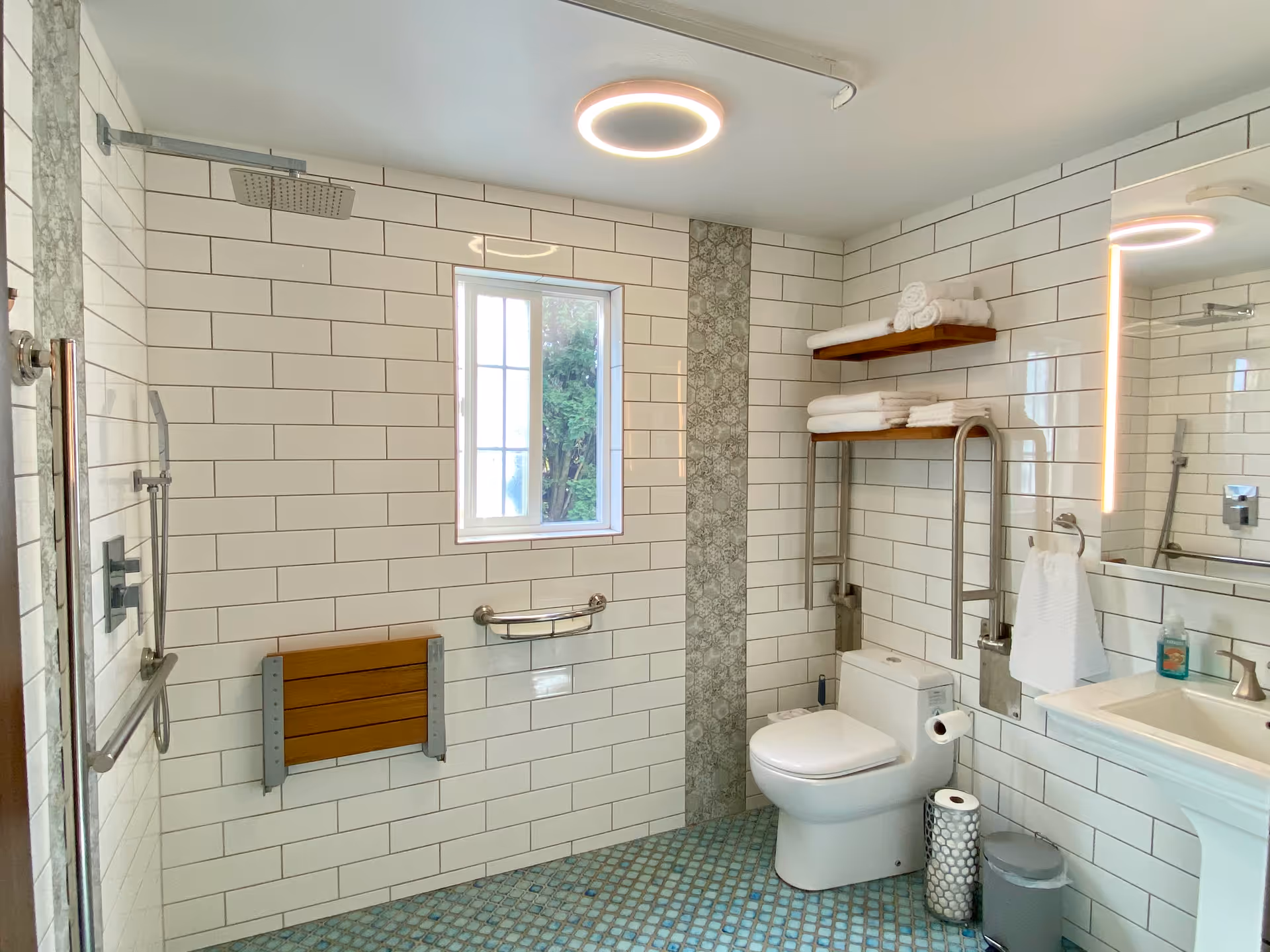 A modern bathroom with white subway tile walls and a blue patterned tile floor. The bathroom features a walk-in shower with a rain showerhead and a fold-down wooden shower seat. There is a window above a grab bar, a white toilet with a toilet paper holder, and a pedestal sink with a towel ring and a lit mirror above it. Two wooden shelves hold neatly folded white towels. A small trash can and a decorative toilet paper holder are on the floor near the toilet.