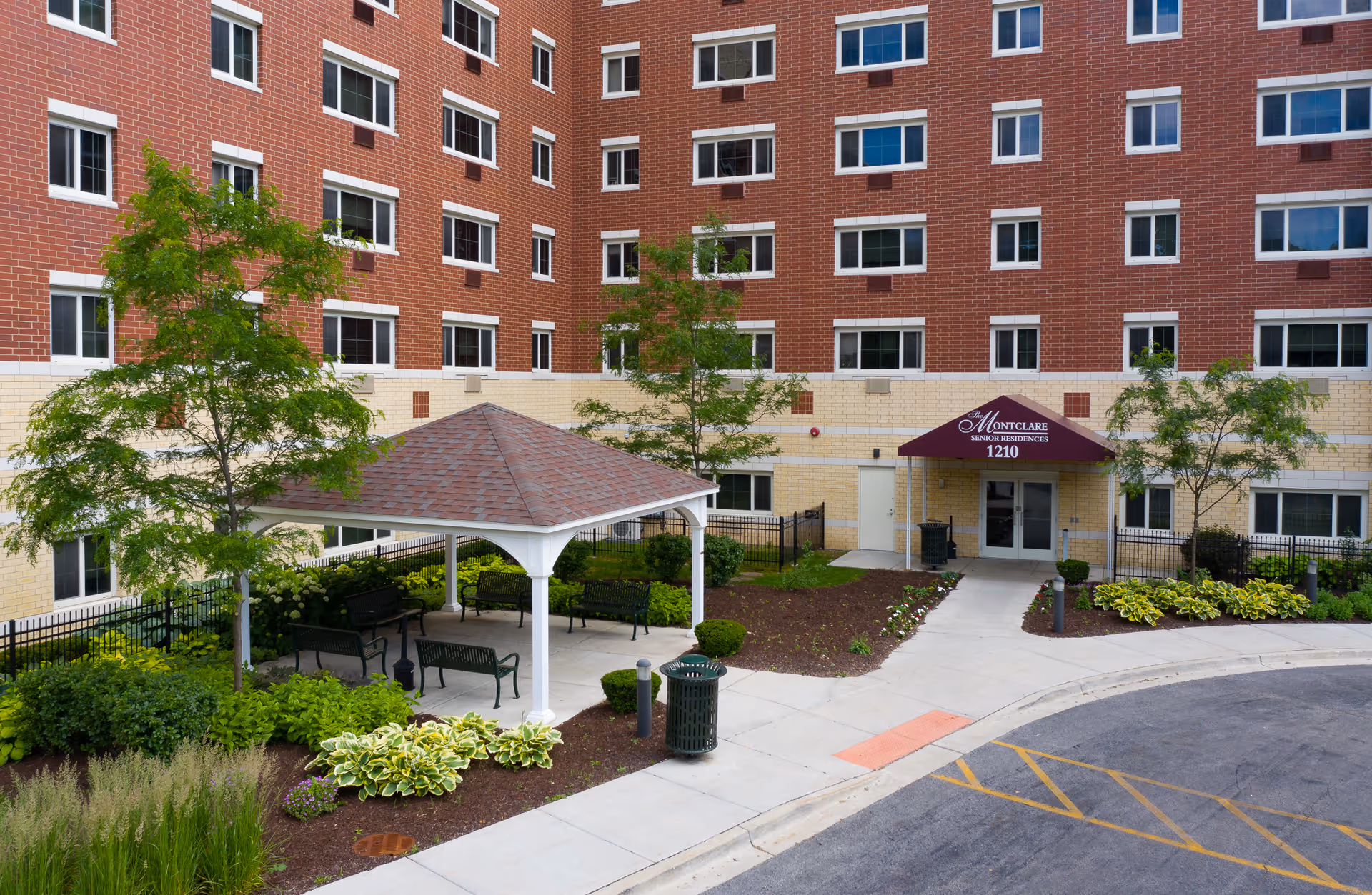 Front entrance of a red-brick senior residence with a gazebo, landscaped courtyard, and an awning reading "Montclare Senior Residences 1210".