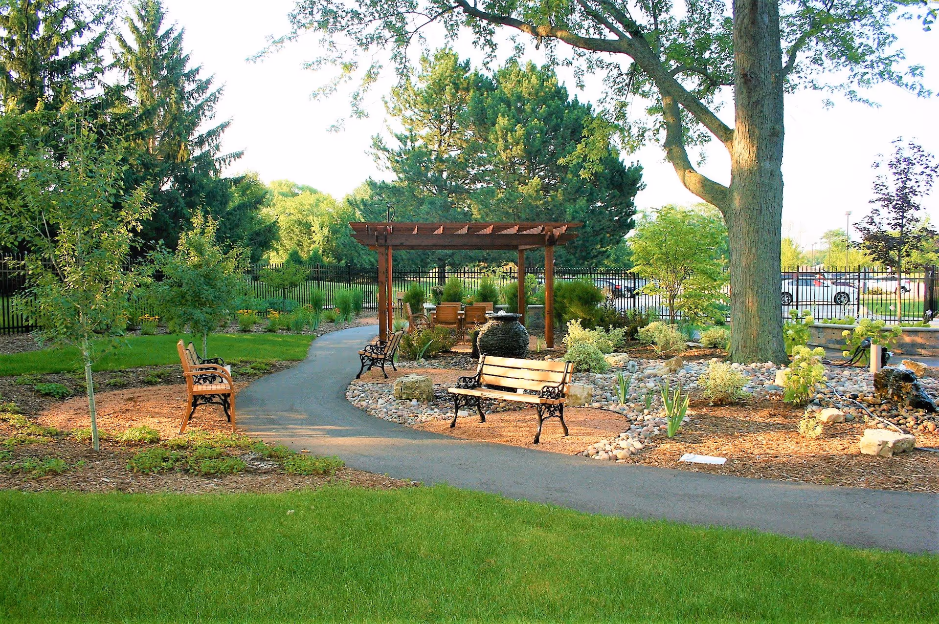 A peaceful outdoor garden area with a paved walking path, wooden benches with black metal armrests, a large tree, various shrubs and plants, and a wooden pergola with seating underneath. A black metal fence and parked cars are visible in the background.