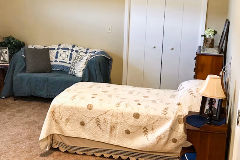 A cozy bedroom featuring a single bed with a floral patterned quilt, a wooden nightstand with a lamp, a wooden dresser with a vase of flowers, and a small couch covered with a blue throw and decorative pillows. There is a closet with double doors in the background.