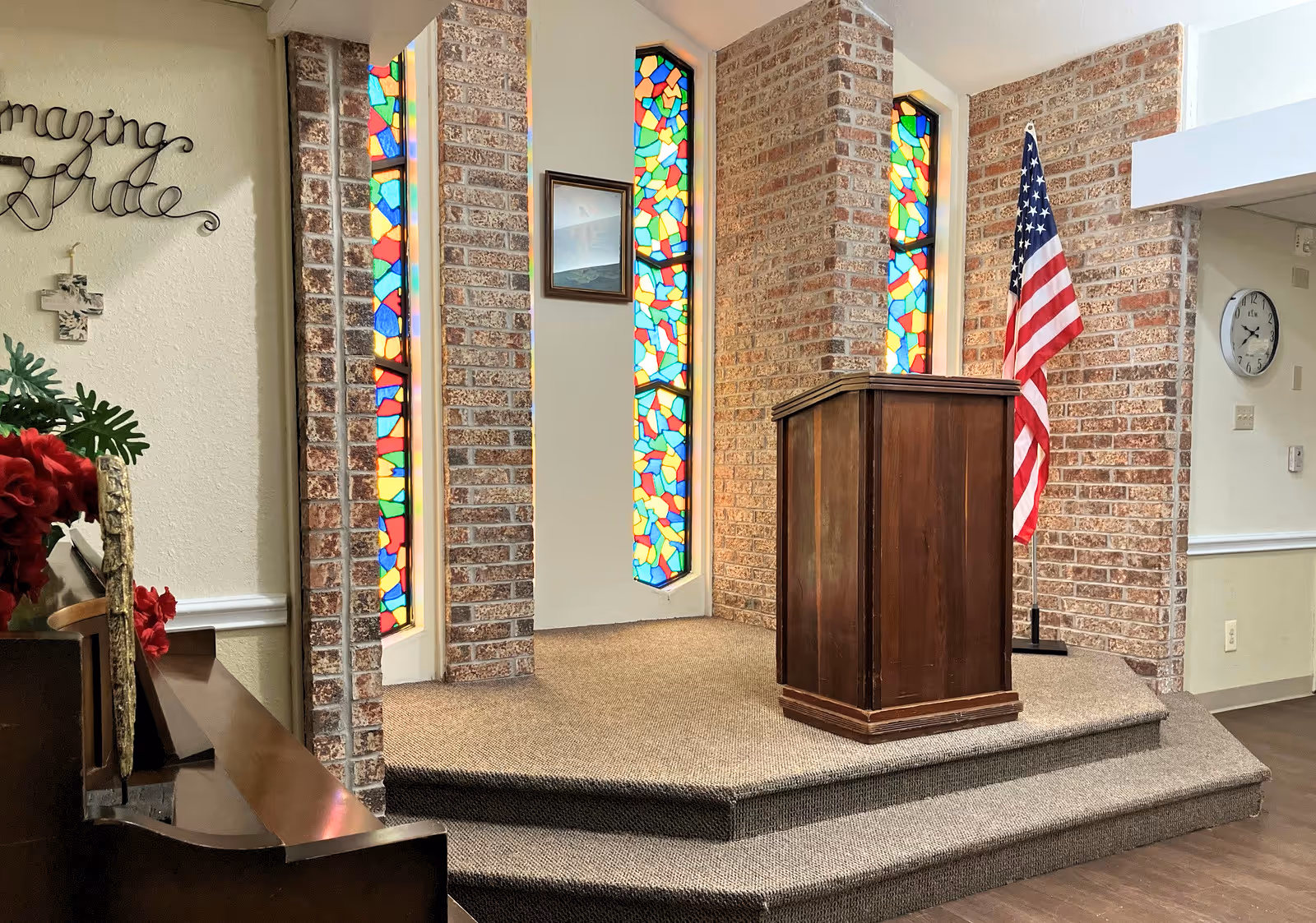Small chapel-style interior with a wooden podium on a raised carpeted platform, stained-glass vertical windows set in a brick wall, and an American flag.