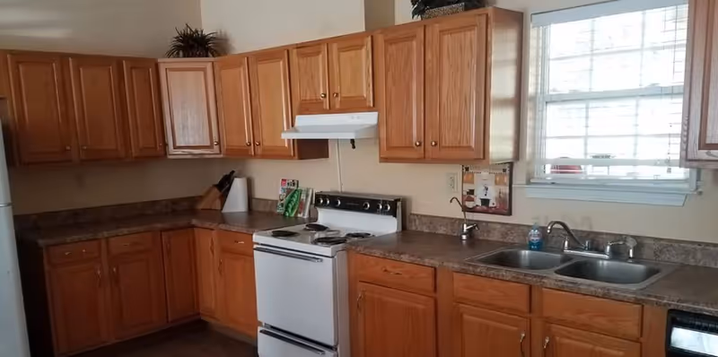 A kitchen with wooden cabinets, a white electric stove with four burners, a double stainless steel sink under a window with blinds, and a countertop with various items including a paper towel holder and a water bottle.