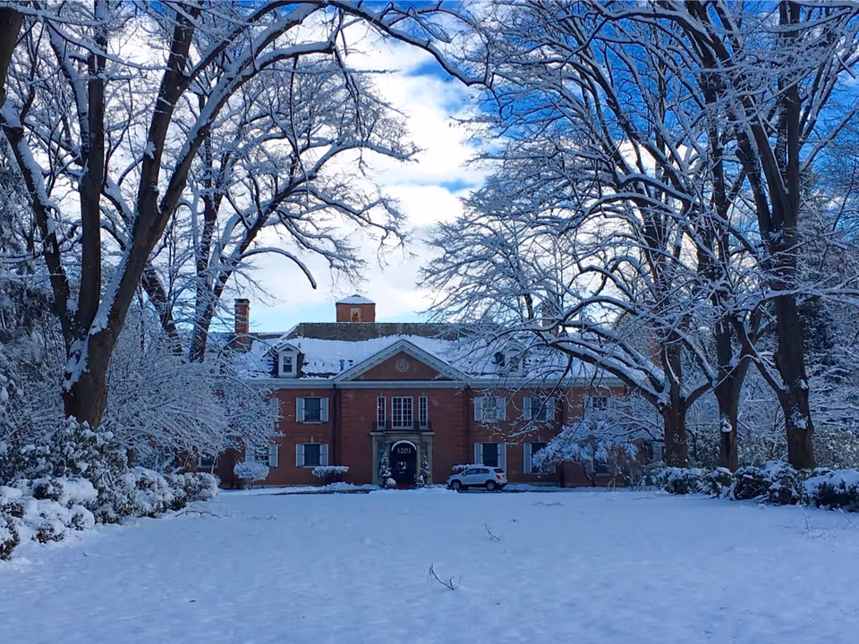 Snow-covered front view of The Villa, a brick senior living building framed by bare trees.