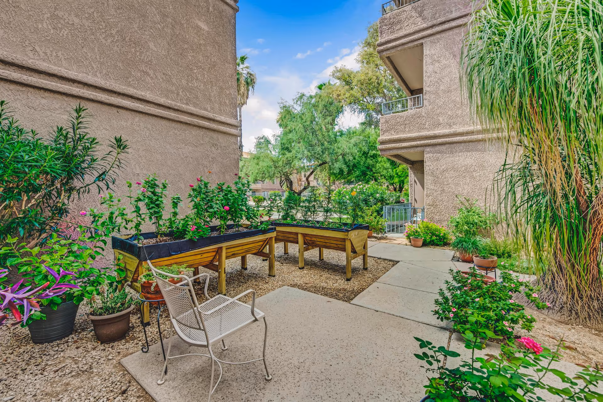 Courtyard garden between stucco buildings featuring raised planter beds, potted plants, a metal chair, and a paved walkway.