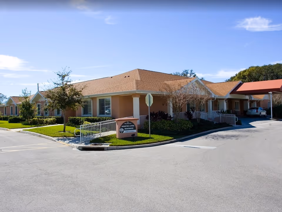 Single-story peach-colored assisted living residence with a covered entrance, ramp, landscaping and driveway under a clear blue sky.