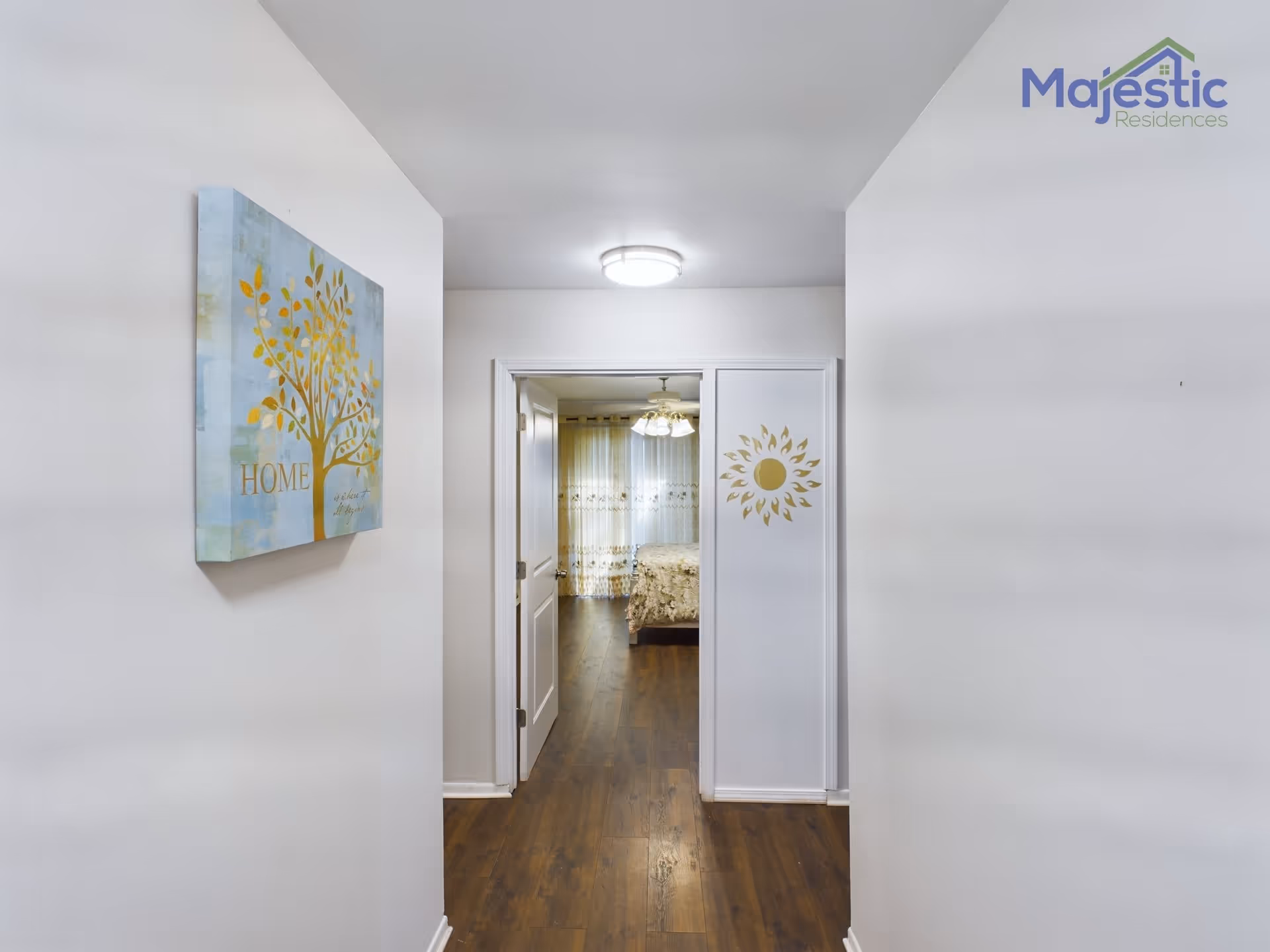 View down a hallway with white walls and wooden flooring leading to a bedroom. On the left wall, there is a decorative painting of a tree with the word 'HOME'. The bedroom at the end of the hallway has a bed with a floral bedspread and sheer curtains with a floral pattern. A ceiling light fixture is visible in the hallway ceiling.