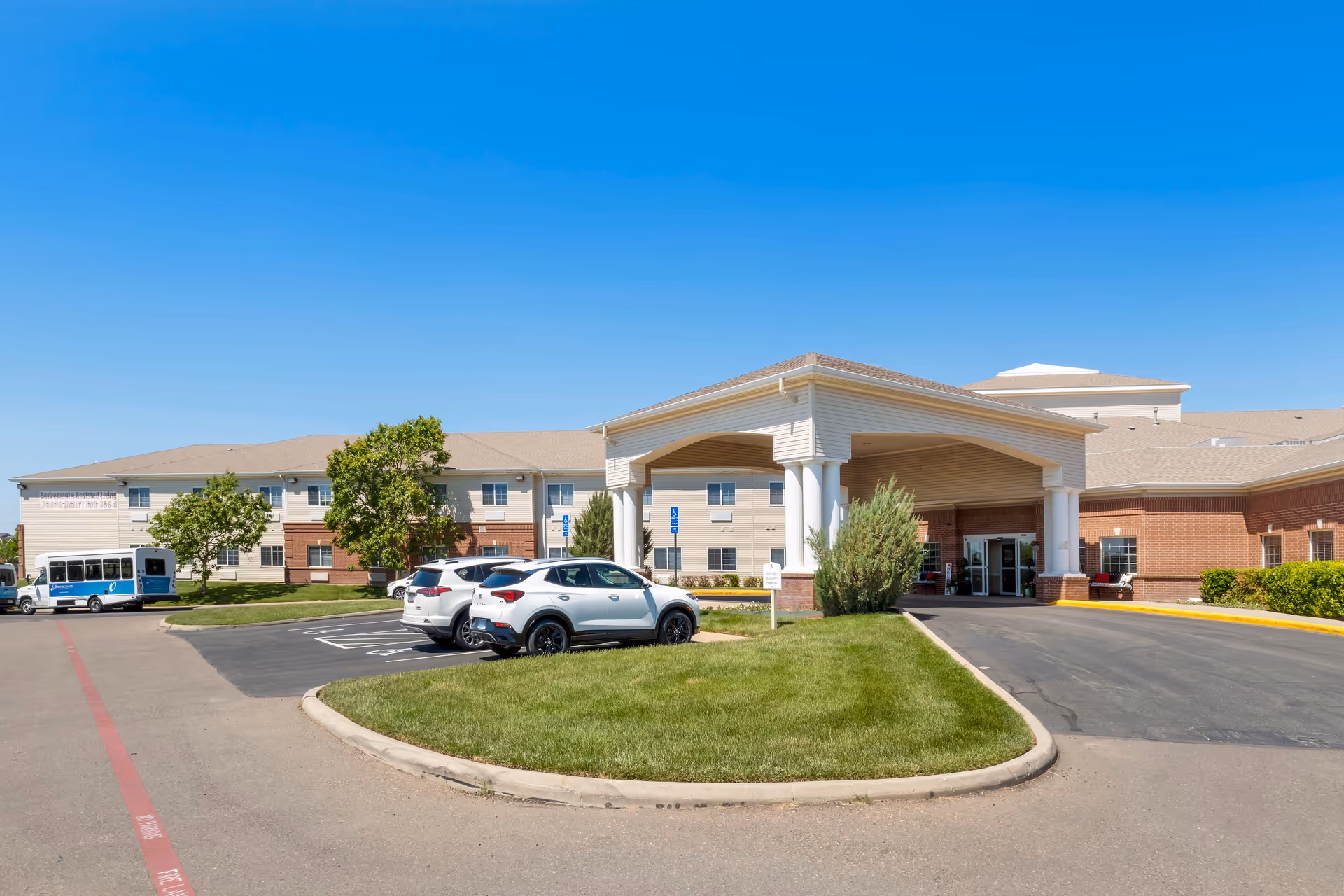 Exterior view of Brookdale Medi Park West facility showing the main entrance with a covered drop-off area, parked cars, a shuttle bus, and a clear blue sky.