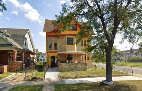 Two-story yellow building with red trim and a front porch on a tree-lined street.