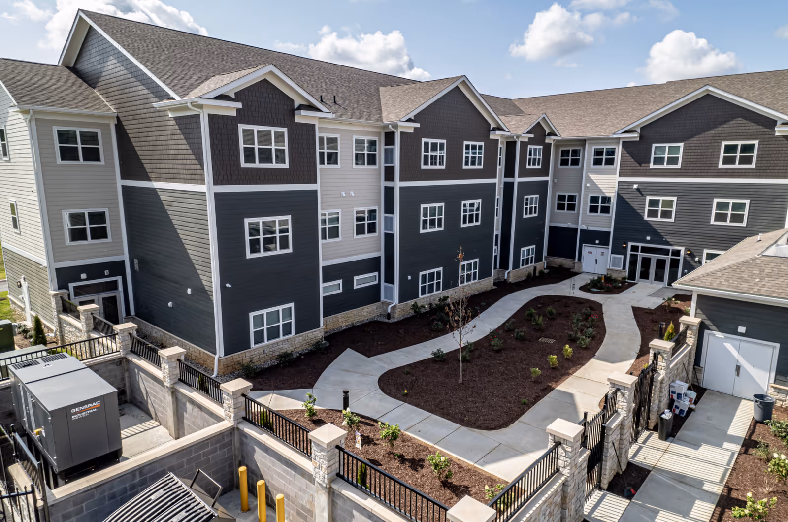 Exterior view of a modern three-story senior living facility building with dark gray and beige siding, multiple windows, and a landscaped courtyard with paved walkways and small plants under a partly cloudy sky.