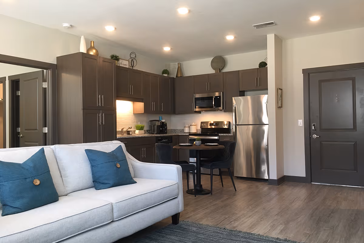 A modern apartment interior featuring a light gray sofa with blue pillows in the foreground. Behind the sofa is a kitchen area with dark brown cabinets, stainless steel appliances including a refrigerator, microwave, and stove. A small round dining table with two chairs is positioned near the kitchen. The floor is wood-style laminate, and the walls are painted light beige. There is a dark-colored door to the right and an open doorway to the left.