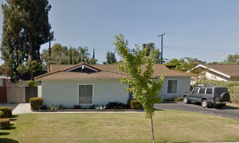 Single-story residential building with a tiled roof, white exterior walls, and a small front yard with grass and a young tree. A dark green SUV is parked on the driveway to the right of the house. Tall trees and other houses are visible in the background under a clear blue sky.
