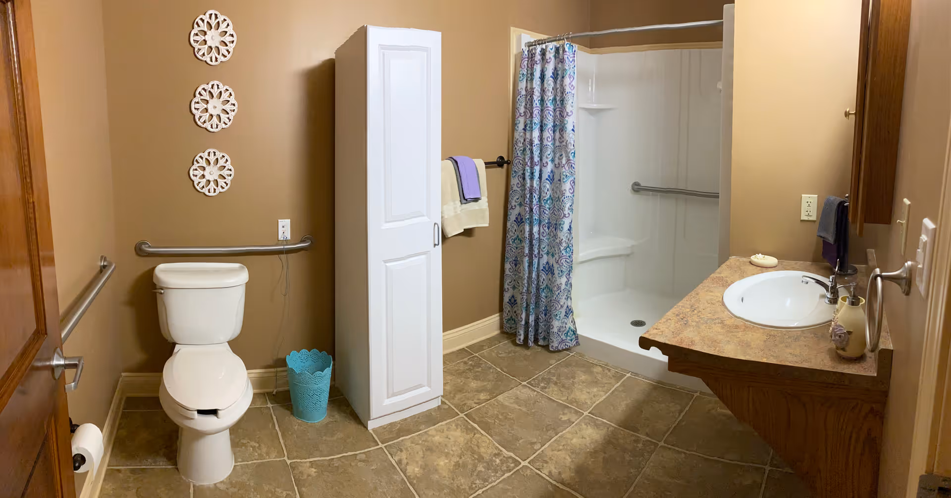 A bathroom with beige walls and tiled floor featuring a white toilet with grab bars on both sides, a white tall storage cabinet, a walk-in shower with a colorful patterned shower curtain, and a wooden vanity with a sink and mirror. There are decorative wall hangings above the toilet and towels hanging on a rack.