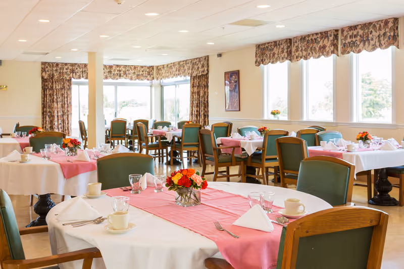 Bright dining room with round tables set with pink runners, floral centerpieces, place settings, and green chairs.