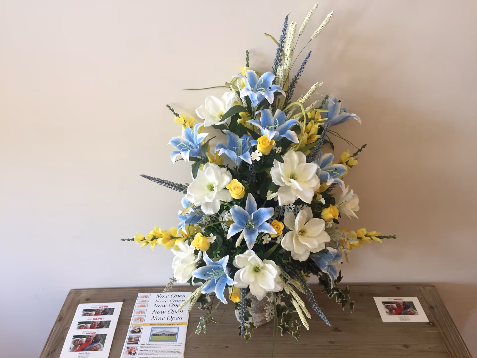 A large floral arrangement with blue, white, and yellow flowers placed on a wooden table. On the table around the arrangement are several flyers and cards, including one that says 'Now Open'. The background is a plain light-colored wall.
