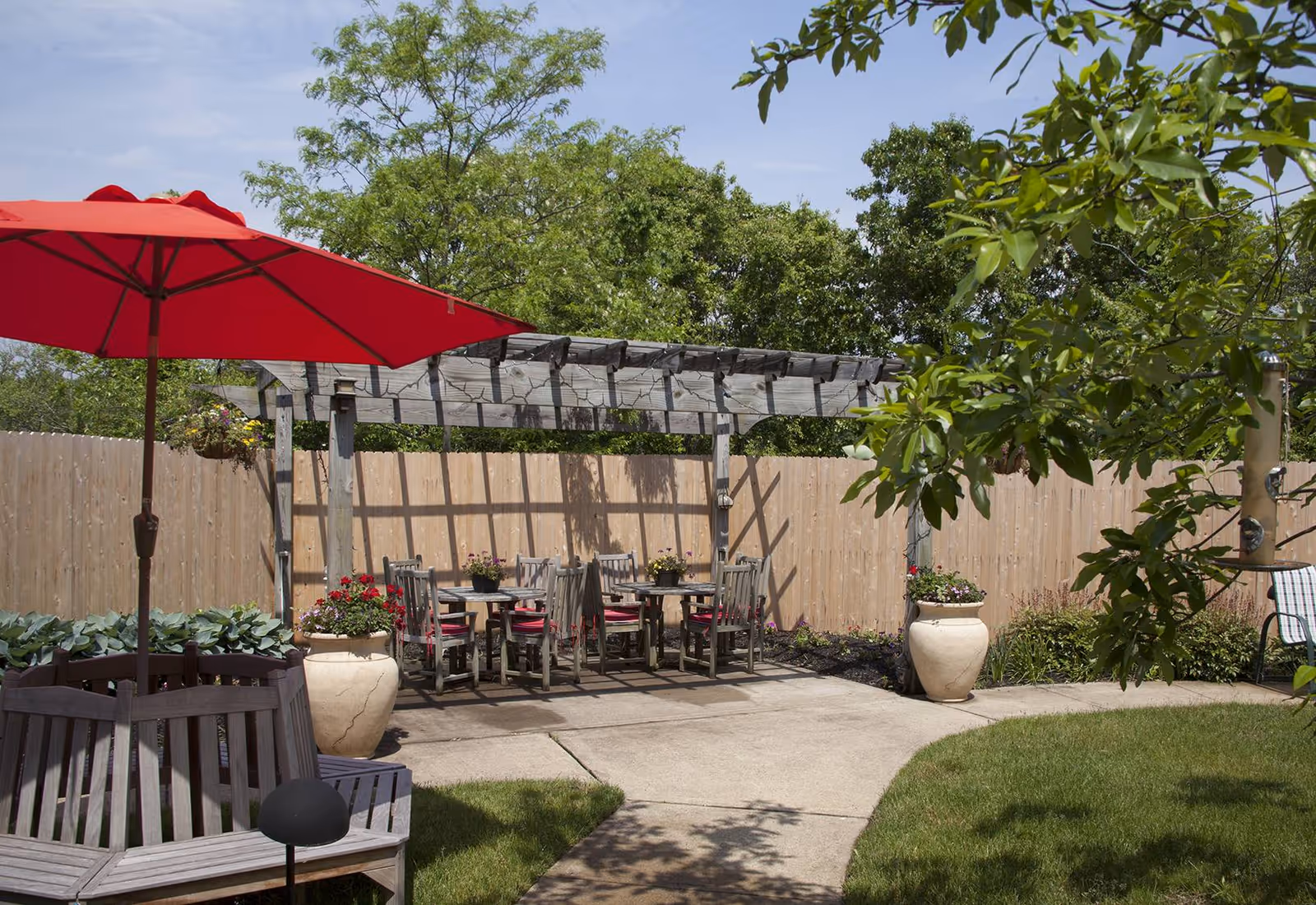 Outdoor patio area with a wooden pergola, several tables and chairs with red cushions, large potted plants with flowers, a red umbrella providing shade, and a wooden bench surrounded by greenery and trees under a clear sky.