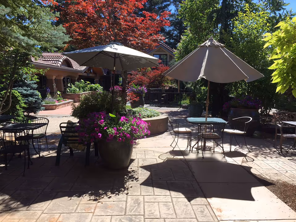 Outdoor patio area with tables and chairs under large umbrellas surrounded by lush greenery and colorful flowers, with a building featuring a tiled roof in the background.