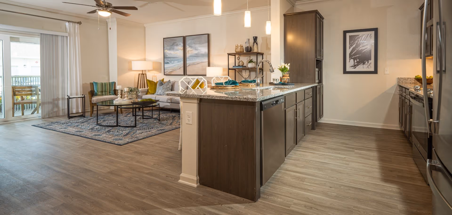 Open-concept living space featuring a modern kitchen with a large island and granite countertops, stainless steel appliances, and dark wood cabinetry. Adjacent living room area with a sofa, armchair, coffee table, decorative pillows, and wall art. Sliding glass doors with white curtains lead to an outdoor balcony with wooden chairs.