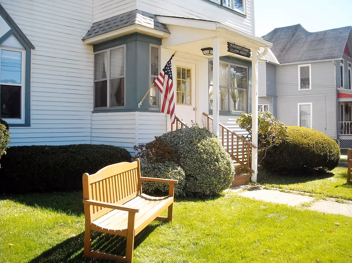 Exterior view of Pinecrest Home For Adults showing a white building with green trim, an American flag mounted near the entrance, a wooden bench on the lawn, and neatly trimmed bushes.