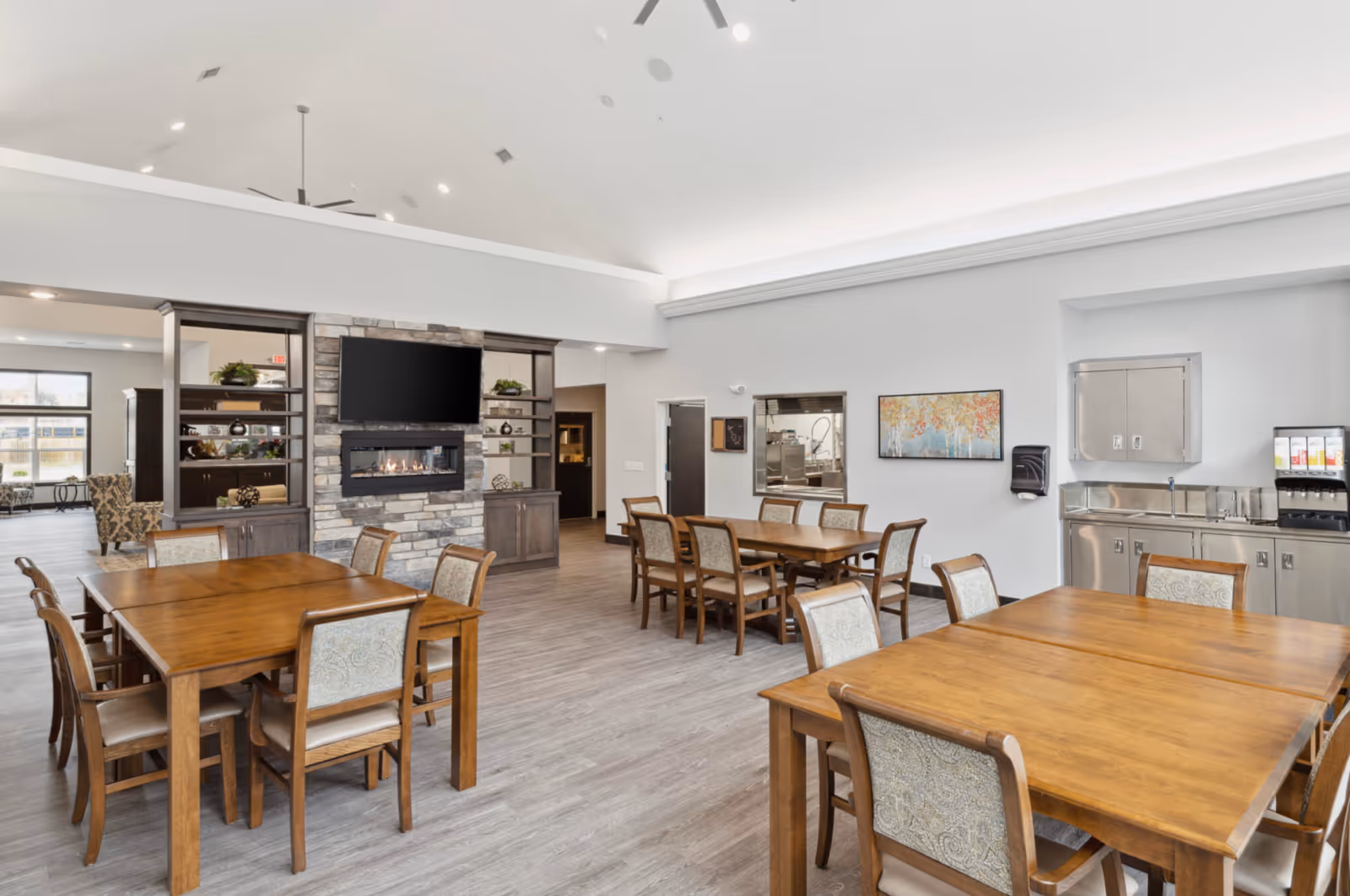 Spacious senior dining room with multiple wooden tables and chairs, a stone fireplace with TV, and a small kitchenette area.